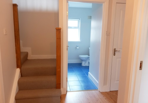 Hallway with stairs and wooden banister leading to blue-tiled bathroom in Leitrim Quay cottage
