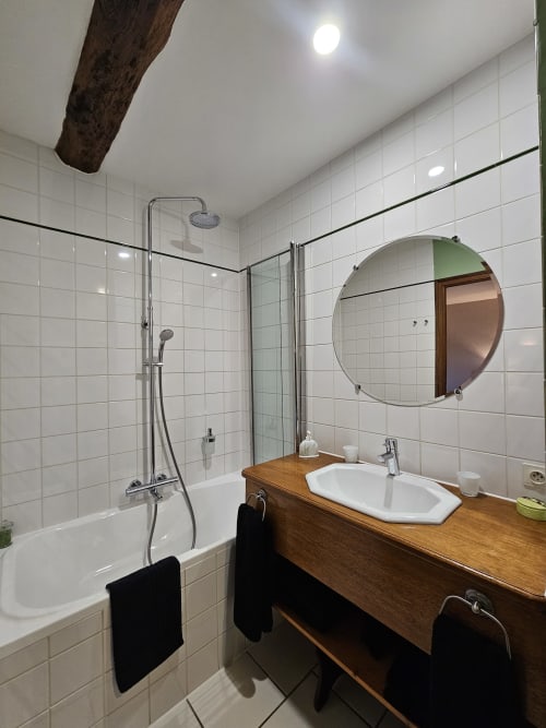 Modern bathroom in Château de Laborde with white tiled walls, round mirror, wooden vanity, glass shower enclosure, and dark
