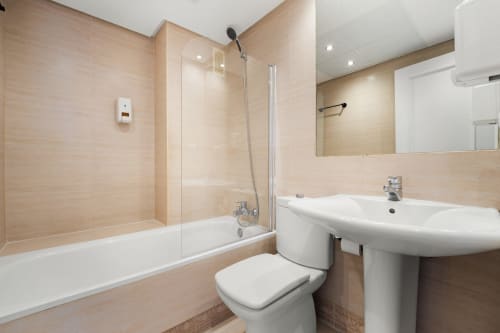 Modern bathroom in Marbella apartment with beige tile walls, white sink, toilet, bathtub and glass shower enclosure