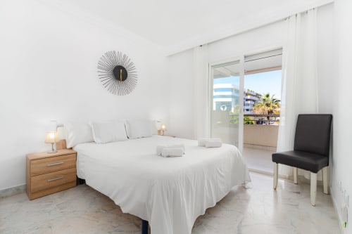 Bright modern bedroom with white bed, wooden nightstand, city view balcony in Marbella apartment