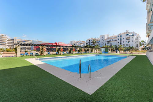 Outdoor swimming pool with blue water, lounge chairs, manicured grass, and Marbella apartment buildings under clear sky