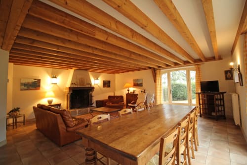 Spacious rustic cottage interior with exposed wooden beams, fireplace, dining table, leather seating and garden views at