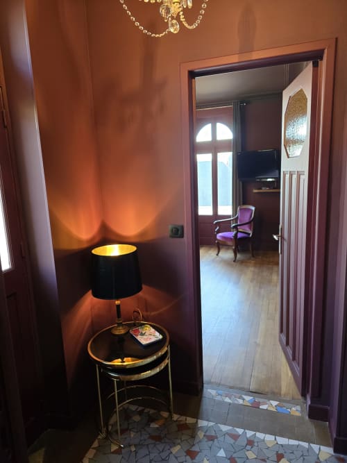 Élégante entrée du couloir du Castel Bayeux B&B avec éclairage chaleureux, lampe noire sur table ronde et porte ouverte sur la chambre