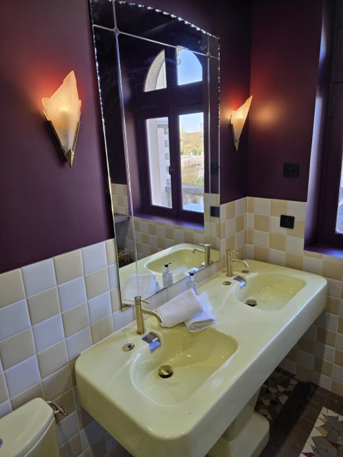 Salle de bain moderne du B&B Le Castel Bayeux avec double vasque, carrelage crème et murs violets ornés d'appliques décoratives