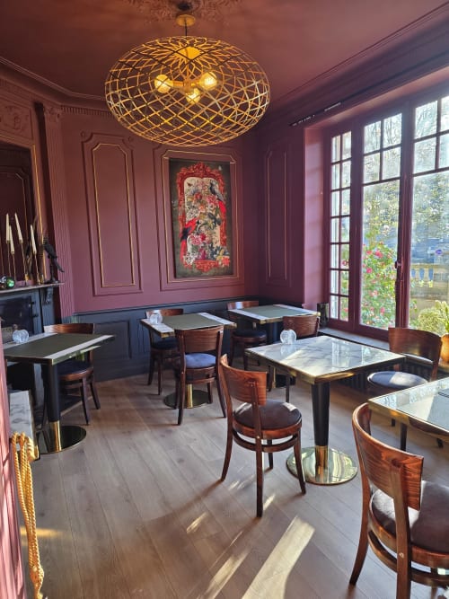 Salle à manger élégante du Castel Bayeux avec murs mauve, luminaire suspendu tressé, tables en bois et vue sur le jardin