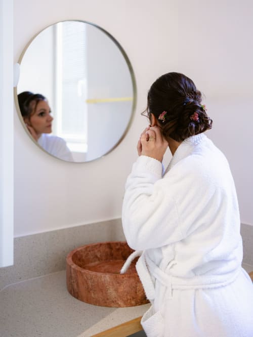 Femme en peignoir blanc se coiffant devant un miroir rond dans la salle de bains de la Maison D'olivier