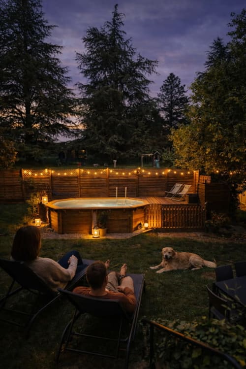 Couple se relaxant près du bain à remous illuminé au crépuscule avec leur chien, gîte Domaine le Manoir en Dordogne