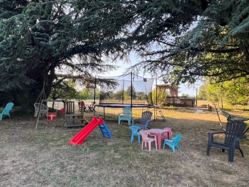 Aire de jeux extérieure du gîte Domaine le Manoir en Dordogne avec toboggan coloré, chaises et trampoline à l'ombre des arbres