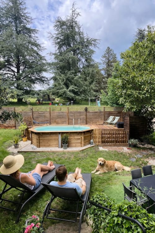 Des clients se détendent au bord de la piscine en bois du Domaine le Manoir à Fossemagne, avec un chien et des transats sur la pelouse