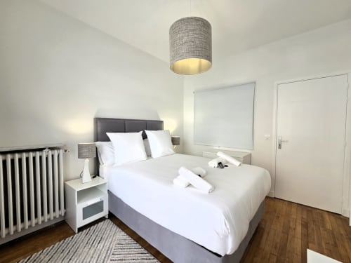Chambre moderne dans un appartement de Levallois-Perret avec lit queen, linge blanc, tête de lit grise, suspension, radiateur et décoration blanche