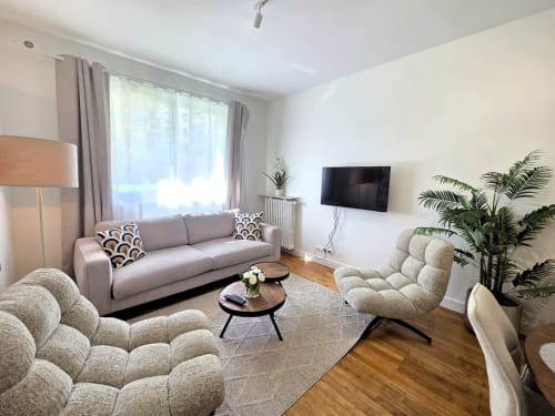 Salon moderne de l'appartement à Levallois-Perret avec canapé gris, fauteuils beige, table en bois, téléviseur, plantes et vue sur le jardin