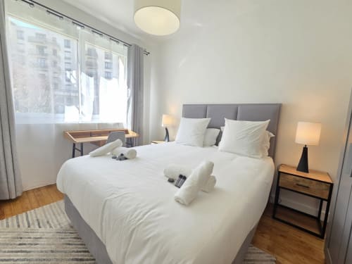 Chambre moderne avec literie blanche, tête de lit grise, bureau en bois et baies vitrées donnant sur les appartements parisiens