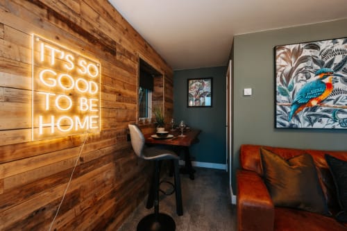 Modern hotel room in Shrewsbury with neon 'It's so good to be home' sign, wooden accent wall, orange sofa, and colorful bird