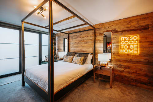 Modern canopy bed in wooden-walled Shrewsbury suite with neon 'It's So Good To Be Home' sign and ensuite bathroom.