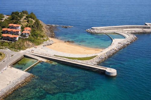 Complejo costero de Mutriku con edificios de tejados naranjas, playa curva y moderno muelle con vistas a aguas turquesas