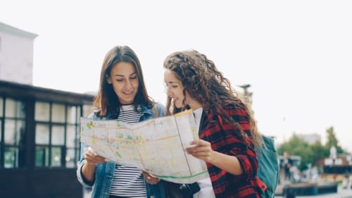 Deux femmes consultant une carte lors d'une exploration à Leysin près de l'Hôtel La Tour d'Aï, avec des bâtiments et des arbres en arrière-plan.