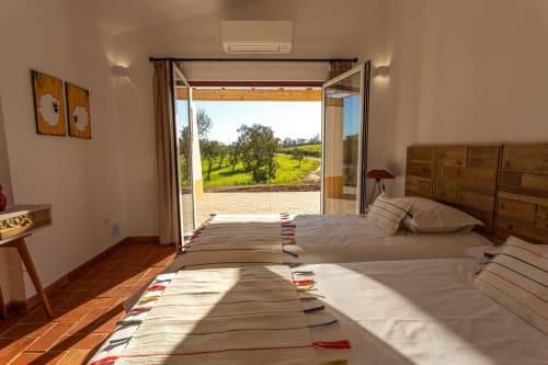 Quarto luminoso com camas de casal, cabeceira em madeira e portas de vidro abertas para olivais em Cerro da Campaniça,