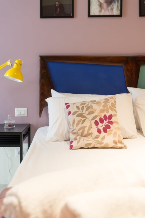 Camera da letto a Tresette Toledo Napoli con biancheria color crema, cuscino floreale, testiera blu, lampada da scrivania gialla e pareti lavanda