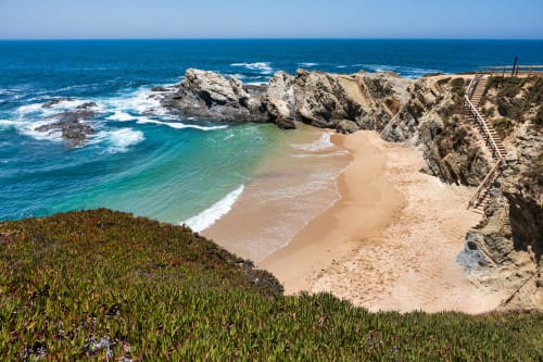 Cove costeira paradisíaca com areia dourada, águas turquesas, penhascos rochosos e escadas de madeira perto de Santiago do Cacém