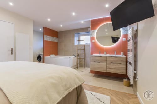 Modern en-suite bedroom with orange and wood accents at La Bergerie De Montitier, featuring illuminated mirror and bathtub
