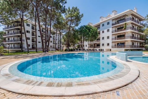 Piscina com bordas curvas no AlgarStays Silves, Portugal, cercada por blocos de apartamentos e pinheiros