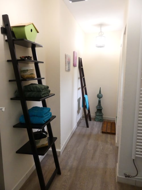 Couloir étroit avec étagère-échelle noire rangement serviettes et décor, parquet, murs blancs, luminaire suspendu à La Cour Des