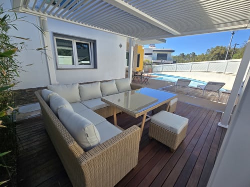 Varanda de villa em Sesimbra com espreguiçadeiras de vime, mesa azul, pérgola branca e vista para a piscina no convés de madeira