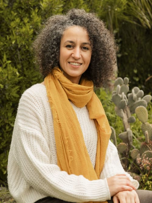 Mujer sonriente en el jardín del hotel Escanda Building Solutions en España, vistiendo suéter crema y bufanda dorada