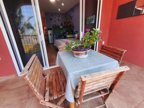 Área de comedor en patio de villa Casa Punta Perula con bancas de madera, macetas con flores, paredes rojas, puertas con vista al océano y cocina