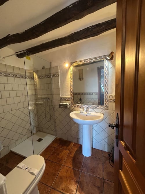 Baño espacioso en la cabaña Casa de la Aguadora con ducha de azulejos blancos, lavabo de pedestal, espejo ornamentado y vigas de madera rústica
