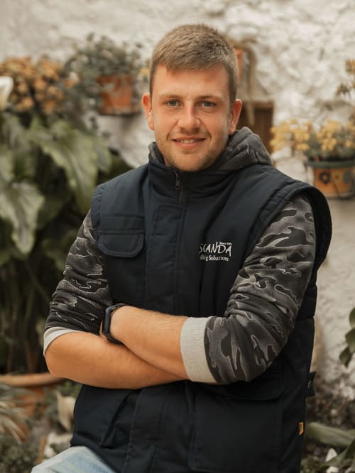 Staff member at Escanda Building Solutions in a greenhouse with potted plants, wearing branded vest and smiling at camera