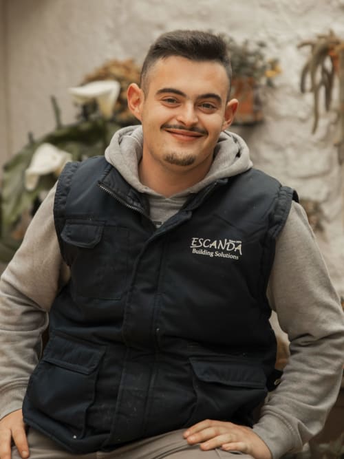 Young man smiling in ESCANDA Building Solutions black vest and gray hoodie, white brick wall background