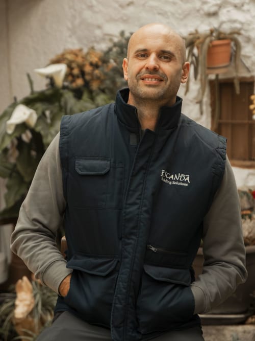 Bald man in Escanda Building Solutions navy vest smiling at rustic courtyard with white walls and plants