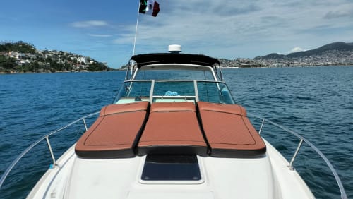 Yate de lujo con tumbonas naranjas amarrado en aguas de Acapulco, bandera mexicana ondeando, colina costera visible