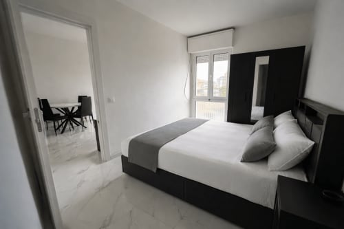 Camera da letto moderna nell'appartamento Anthea Loft Elegans 2 a Lignano Sabbiadoro, con letto nero, biancheria bianca, vista sulla città e zona pranzo