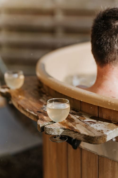 Client savourant un verre de vin blanc dans une baignoire en bois lors d'une expérience spa aux Granges De Pil Bara, Langoat