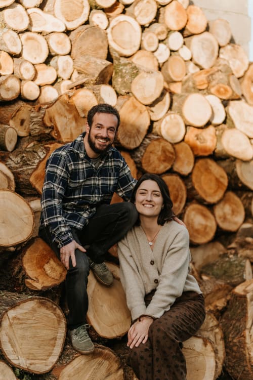Couple souriant aux Granges de Pil Bara à Langoat, France, posant devant du bois de chauffage empilé