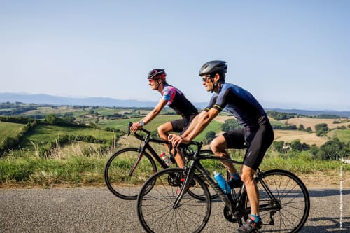 Deux cyclistes roulent sur une route pittoresque avec des collines ondulantes et des montagnes en arrière-plan près d'Ax-les-Thermes