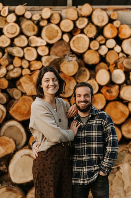 Couple souriant aux Granges De Pil Bara, gîte en Langoat, France, avec du bois de chauffage empilé en arrière-plan