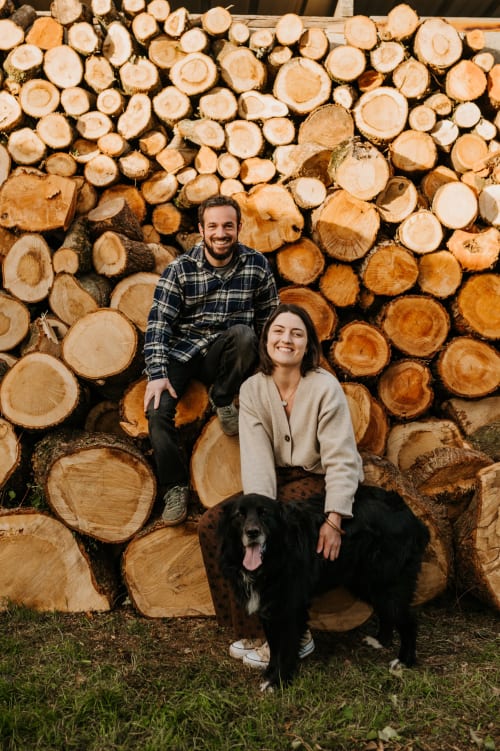 Couple avec chien noir posant devant des bûches empilées aux Granges de Pil Bara, un charmant gîte à Langoat, France