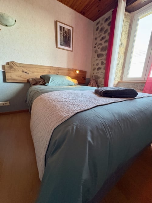 Gemütliches Schlafzimmer in der Pension Lo Soulenquo in Golinhac mit Holzkopfteil, blauen Kissen und Bergblick durchs Fenster