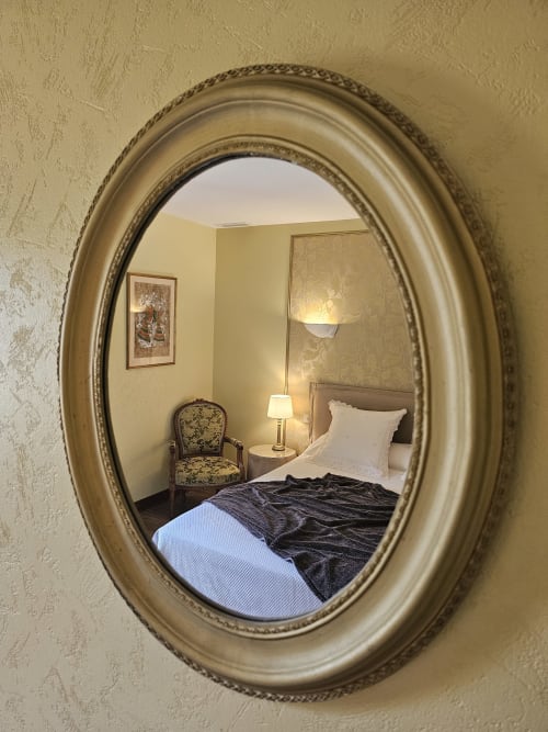 Miroir ovale orné reflétant la chambre du Château de Ribourdin, avec lit, fauteuil et table de nuit avec lampe