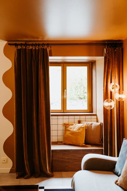 Coin douillet aux Granges De Pil Bara avec rideaux terracotta, banquette fenêtre garnie de coussins et suspensions