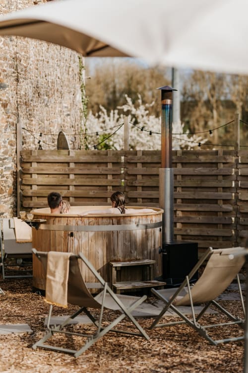 Couple se relaxant dans un bain à remous en bois au gîte Les Granges De Pil Bara à Langoat, France, avec vue sur la forêt et salon de patio.