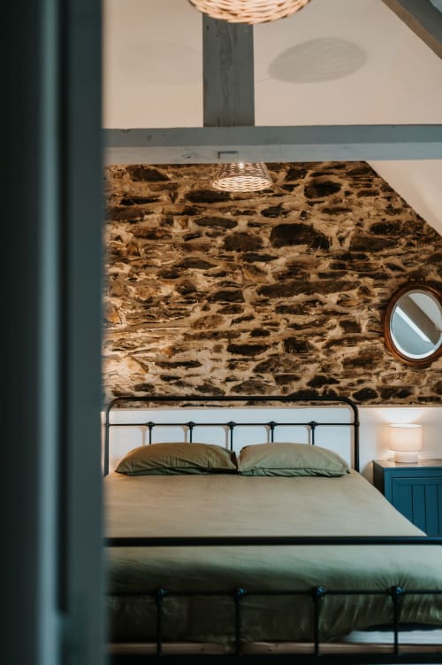 Chambre cosy du gîte Les Granges De Pil Bara avec mur en pierre, lit en métal, coussins verts et décor breton rustique