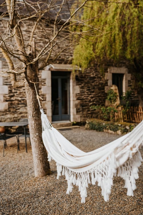 Hamac blanc à franges suspendu entre un arbre nu et un cottage en pierre aux Granges De Pil Bara, Langoat
