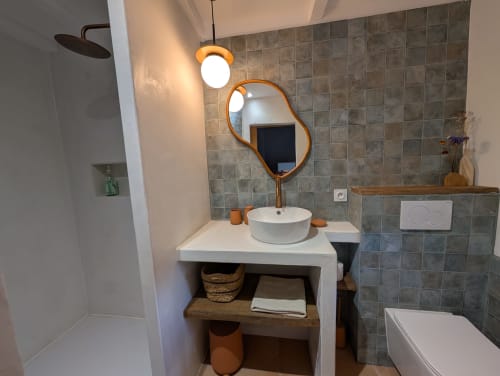 Modern bathroom at Domaine de Pelican with round white sink, organic curved mirror, tiled walls, and wooden vanity shelf with