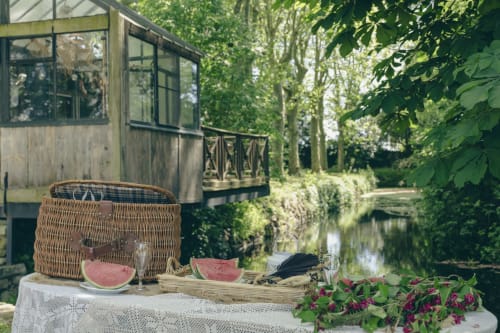 Installation pique-nique au cottage Domaine ForRest à Lay, France avec panier en osier, pastèque et fleurs au bord d'un canal