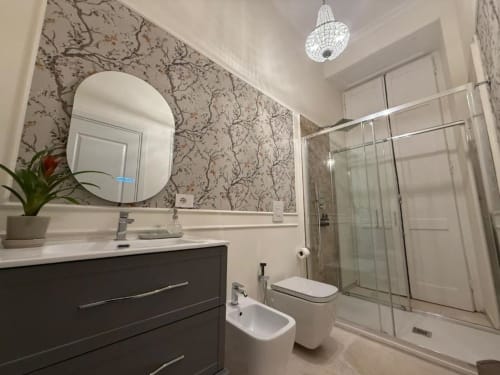 Moderno bagno della Residenza Seconnino Napoli con vanità scura, specchio rotondo, carta da parati decorata e box doccia in vetro