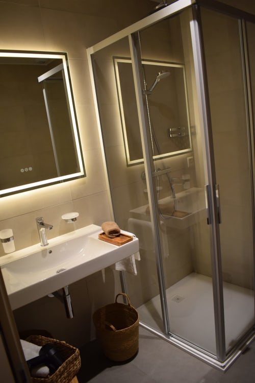 Salle de bains moderne du Boutique Hôtel La Couronne avec miroir lumineux, vasque blanche, douche vitrée et rangements tissés
